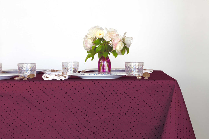 Staccato Nero shibori 100% cotton tablecloth in deep cerise pink on table with full place settings, mosaic garden dinner plates, hand-painted confetti glasses, and a hand-painted vase with roses against a white background 