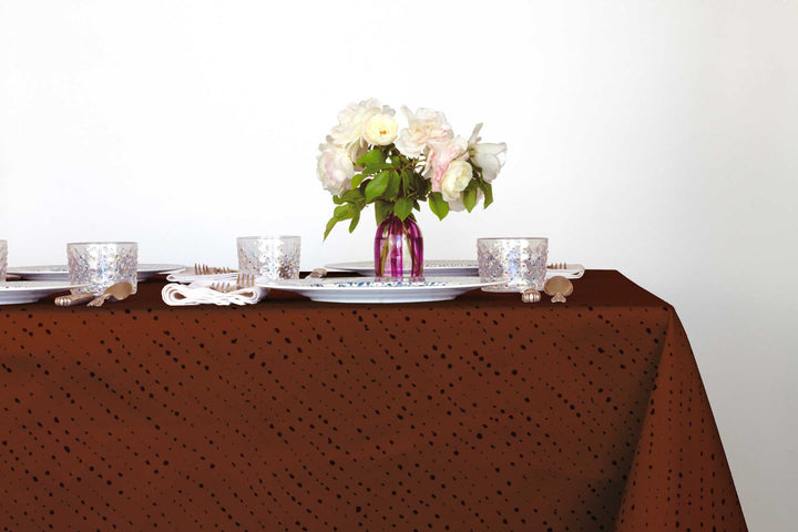Staccato Nero shibori 100% cotton tablecloth in rich russet brown on table with full place settings, mosaic garden dinner plates, hand-painted confetti glasses, and a hand-painted vase with roses against a white background
