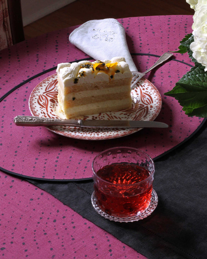 Venable Moore tabletop collection made to order Staccato Nero Shibori Cerise pink tablecloth and placemat with Mosaic garden plate in chestnut with hand-painted one-of-a-kind glass in lapis blue on table with flowers and slice of cake 