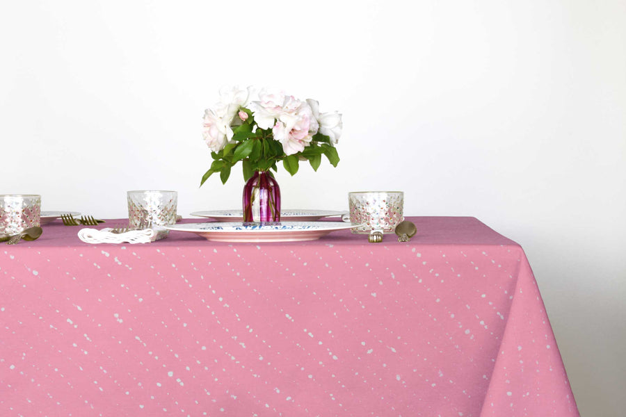 Staccato sbiancato shibori 100% cotton tablecloth in fresh carnation pink on table with full place settings, mosaic garden dinner plates, hand-painted confetti glasses, and a hand-painted vase with roses against a white background