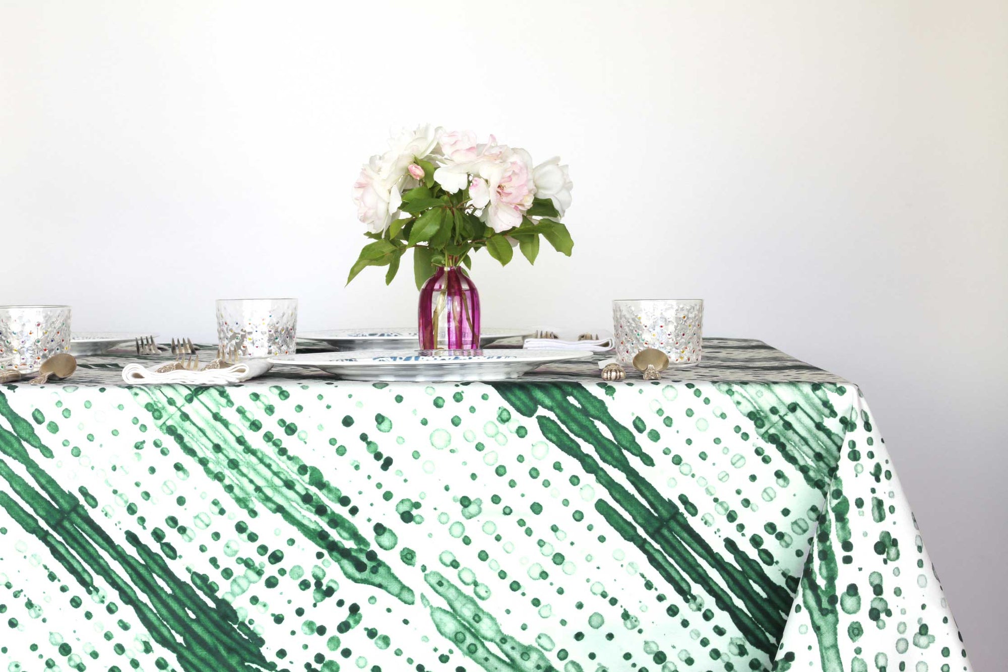 Glissando shibori 100% cotton tablecloth in verdant emerald green on table with full place settings, mosaic garden dinner plates, hand-painted confetti glasses, and a hand-painted vase with roses against a white background 