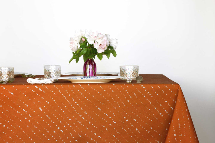 Staccato sbiancato shibori 100% cotton tablecloth in earthy ginger orange on table with full place settings, mosaic garden dinner plates, hand-painted confetti glasses, and a hand-painted vase with roses against a white background
