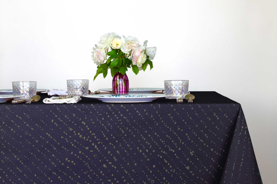 Staccato decolorato shibori 100% cotton tablecloth in inky midnight blue on table with full place settings, mosaic garden dinner plates, hand-painted confetti glasses, and a hand-painted vase with roses against a white background 