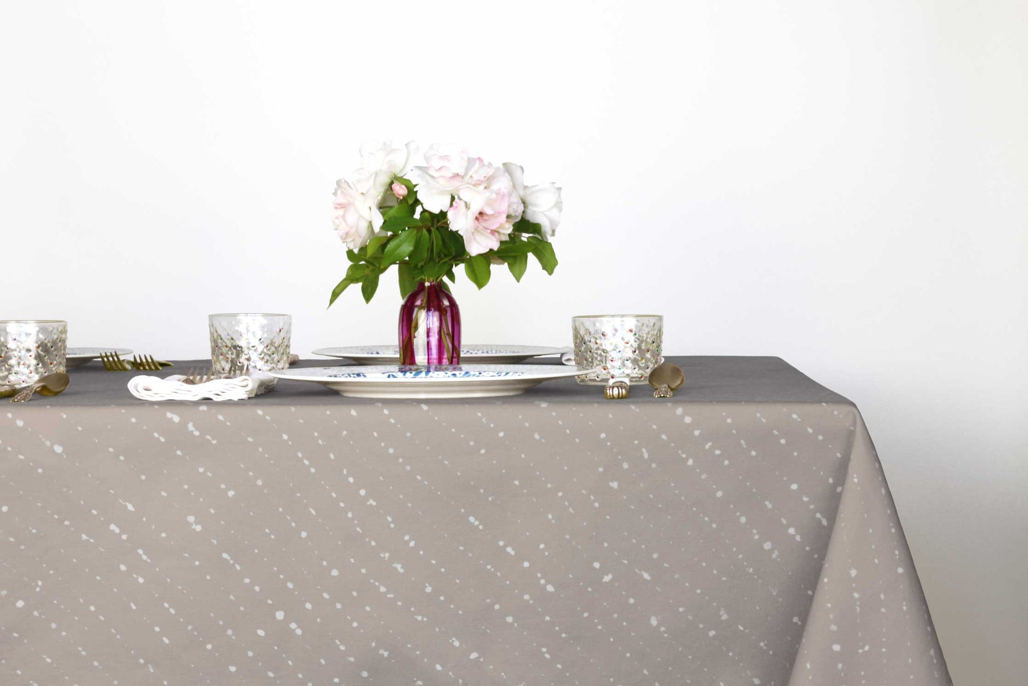  Staccato sbiancato shibori 100% cotton tablecloth in neutral flax tan on table with full place settings, mosaic garden dinner plates, hand-painted confetti glasses, and a hand-painted vase with roses against a white background