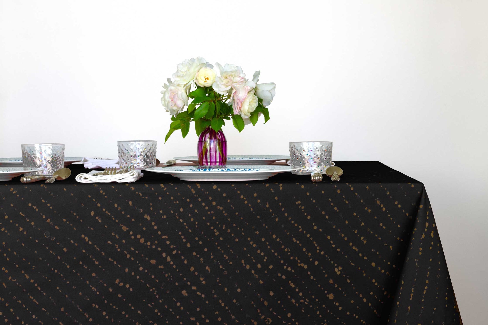 Venable Moore Staccato decolorato shibori 100% cotton tablecloth in onyx black on table with full place settings, mosaic garden dinner plates, hand-painted confetti glasses, and a hand-painted vase with roses against a white background 
