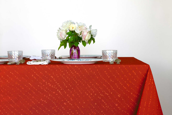 Staccato decolorato shibori 100% cotton tablecloth in vibrant paprika red on table with full place settings, mosaic garden dinner plates, hand-painted confetti glasses, and a hand-painted vase with roses against a white background 