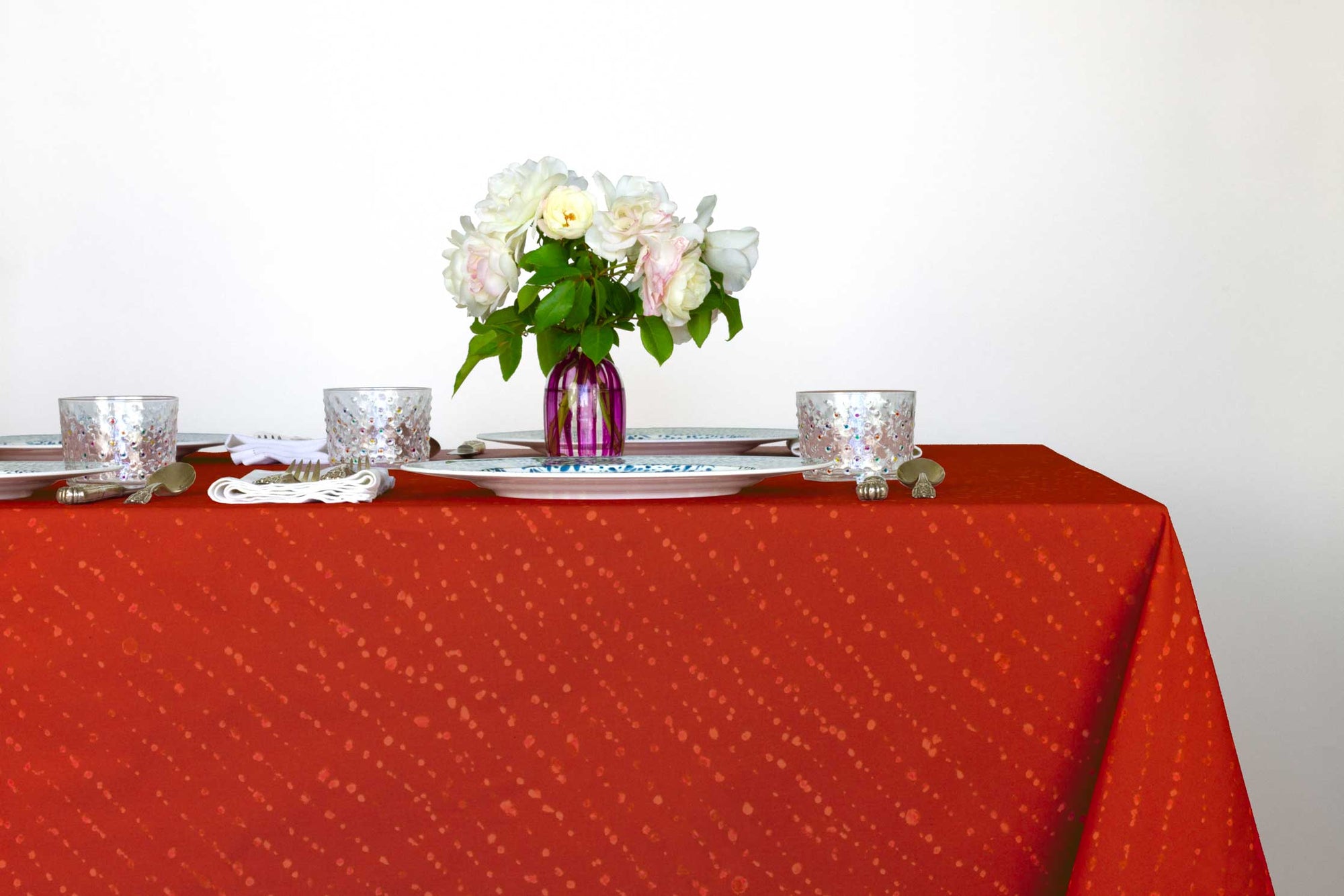 Venable Moore Staccato decolorato shibori 100% cotton tablecloth in vibrant paprika red on table with full place settings, mosaic garden dinner plates, hand-painted confetti glasses, and a hand-painted vase with roses against a white background 