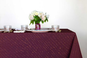 Staccato decolorato shibori 100% cotton tablecloth in saturated plum purple on table with full place settings, mosaic garden dinner plates, hand-painted confetti glasses, and a hand-painted vase with roses against a white background 