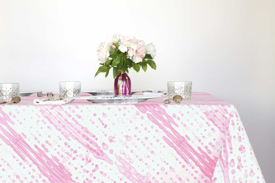 Glissando shibori 100% cotton tablecloth in delicate posy pink on table with full place settings, mosaic garden dinner plates, hand-painted confetti glasses, and a hand-painted vase with roses against a white background 