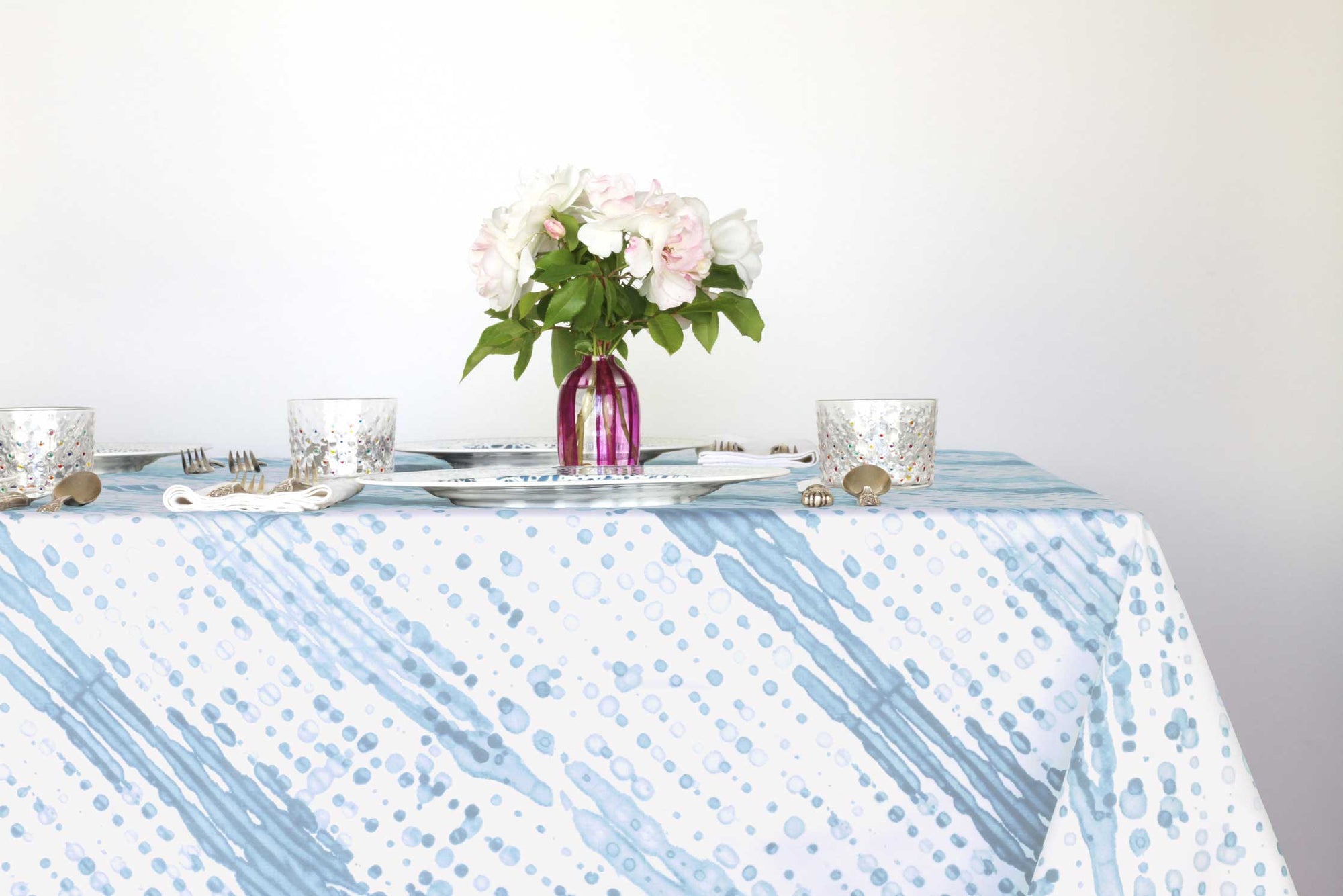 Glissando shibori 100% cotton tablecloth in soft powder blue on table with full place settings, mosaic garden dinner plates, hand-painted confetti glasses, and a hand-painted vase with roses against a white background 