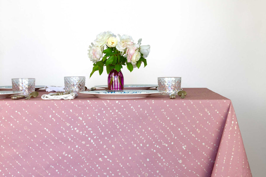 Staccato decolorato shibori 100% cotton tablecloth in dusty rose clay pink on table with full place settings, mosaic garden dinner plates, hand-painted confetti glasses, and a hand-painted vase with roses against a white background 