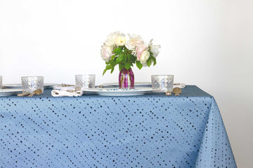 Staccato Nero shibori 100% cotton tablecloth in clear sky blue on table with full place settings, mosaic garden dinner plates, hand-painted confetti glasses, and a hand-painted vase with roses against a white background 