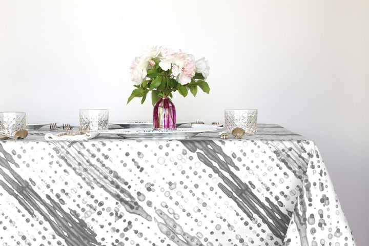 Glissando shibori 100% cotton tablecloth in neutral storm grey on table with full place settings, mosaic garden dinner plates, hand-painted confetti glasses, and a hand-painted vase with roses against a white background 