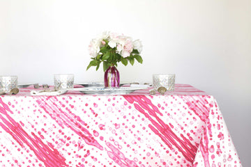 Glissando shibori 100% cotton tablecloth in juicy strawberry pink on table with full place settings, mosaic garden dinner plates, hand-painted confetti glasses, and a hand-painted vase with roses against a white background