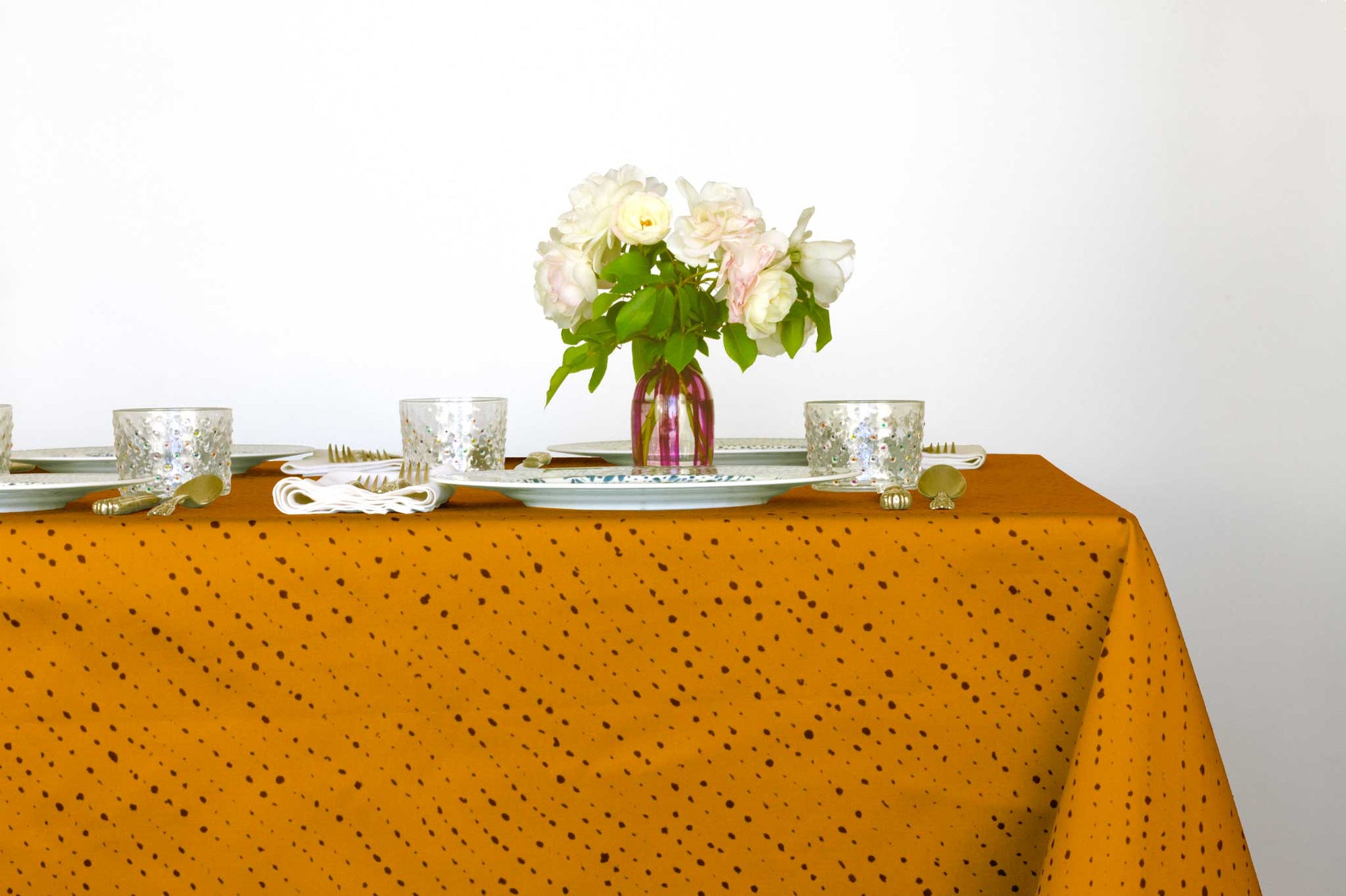 Venable Moore Staccato Nero shibori 100% cotton tablecloth in bold marigold yellow on table with full place settings, mosaic garden dinner plates, hand-painted confetti glasses, and a hand-painted vase with roses against a white background
