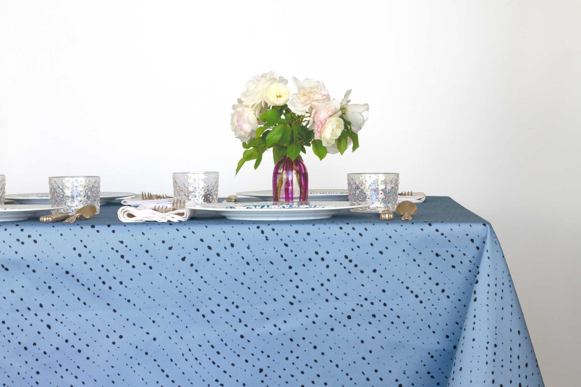 Venable Moore Staccato Nero shibori 100% cotton tablecloth in clear sky blue on table with full place settings, mosaic garden dinner plates, hand-painted confetti glasses, and a hand-painted vase with roses against a white background 
