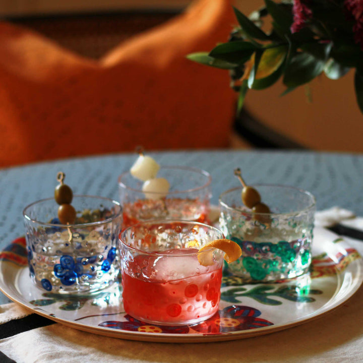 Venable Moore Hand-painted one-of-a-kind Bubble glasses in tomato red, tangerine orange, shamrock green, and lapis blue filled with cocktails on a tray on a table in cozy light