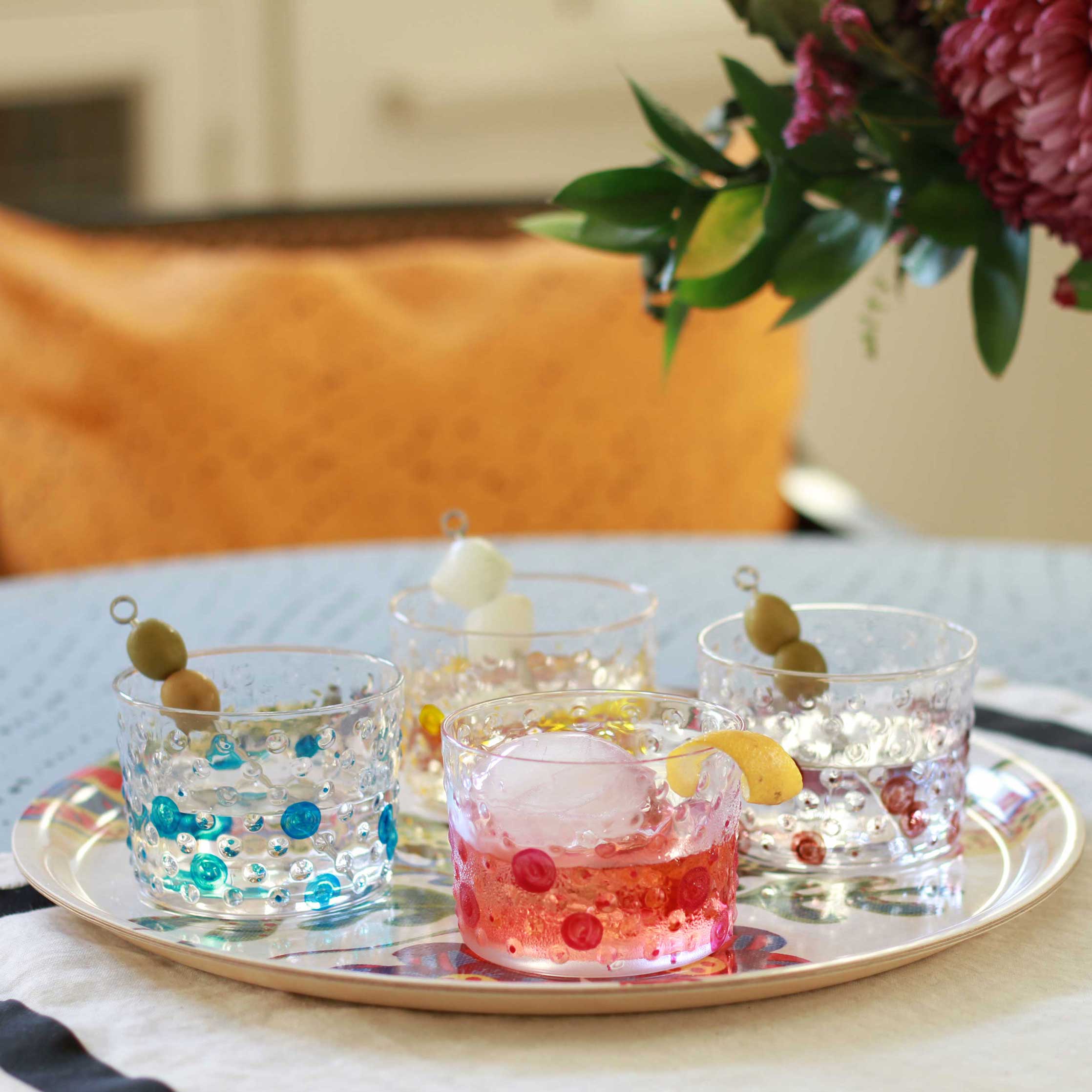Venable Moore hand-painted one-of-a-kind Bubble glasses in turqoise blue, magenta pink, sun yellow, and coffee brown filled with cockatils on tray on table with flowers 