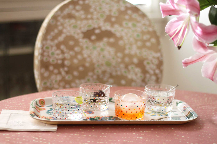 venable moore hand-painted made-to-order confetti glasses with cocktails on a tray on venable moore staccato decolorato shibori tablecloth in rose clay wtih venable moore frizzante linen in  buff on chair with lillies on the side