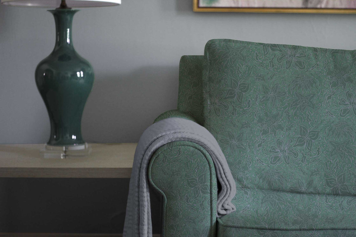 Fiori hand drawn floral cotton in teal green upholstered on a sofa with a side table and lamp and installed in a living room