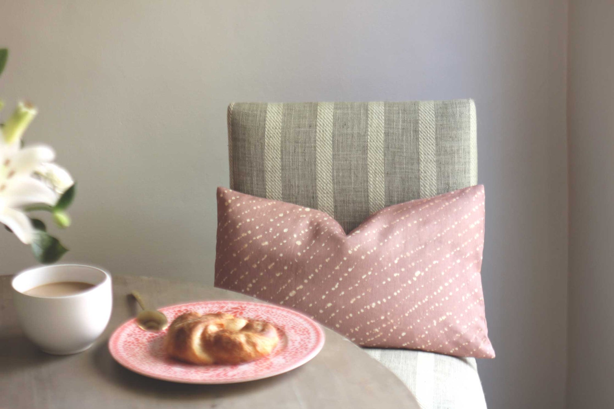 'Staccato Decolorato' Shibori linen pillow in Rose Clay pink on chair with table, Mosaic garden porcelain plate in orchid with croissant, and cup of coffee